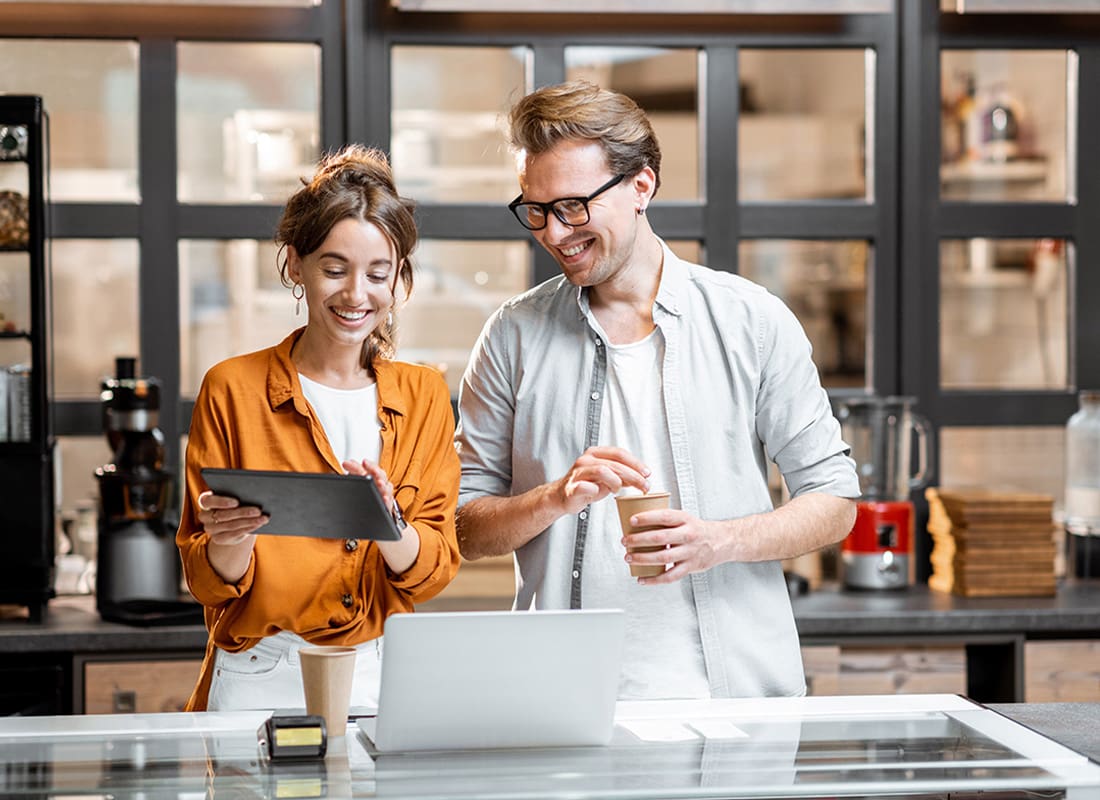 Business Insurance - Cheerful Young Female and Male Small Business Owners Standing Behind the Front Counter of Their Cafe Looking at a Tablet